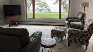 Un salon avec une grande fenêtre, un téléviseur posé sur un meuble, un canapé, un fauteuil et une petite table.