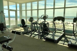A gym with cardio machines and dumbbells, large windows with a beach view.