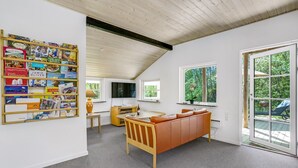 Una sala de estar acogedora con techo de madera, una estantería llena de libros, un sofá de cuero marrón y una zona de comedor con mesa y sillas.