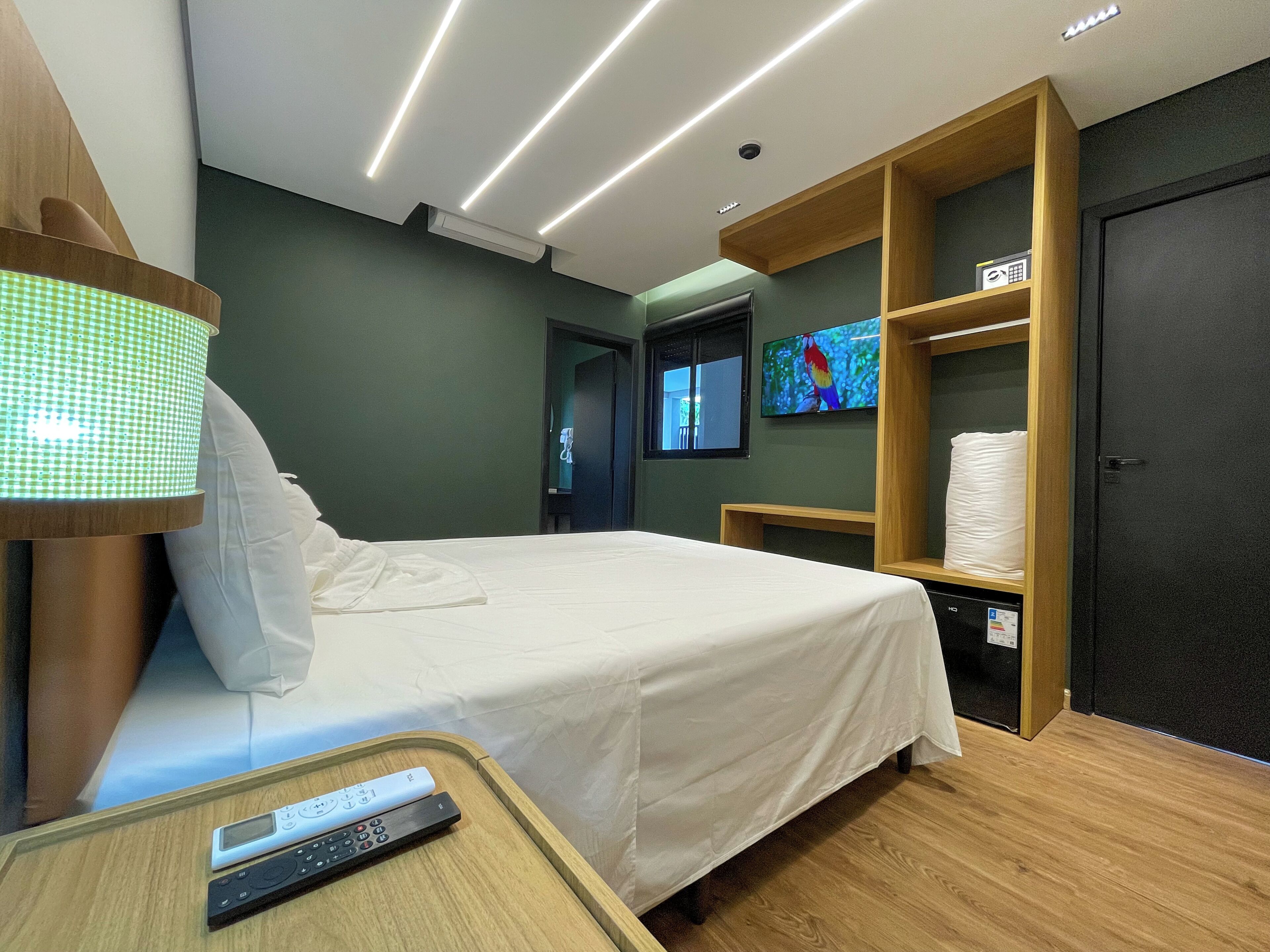 A modern hotel room with a bed, bedside lamp, TV, and a shelf with towels.
