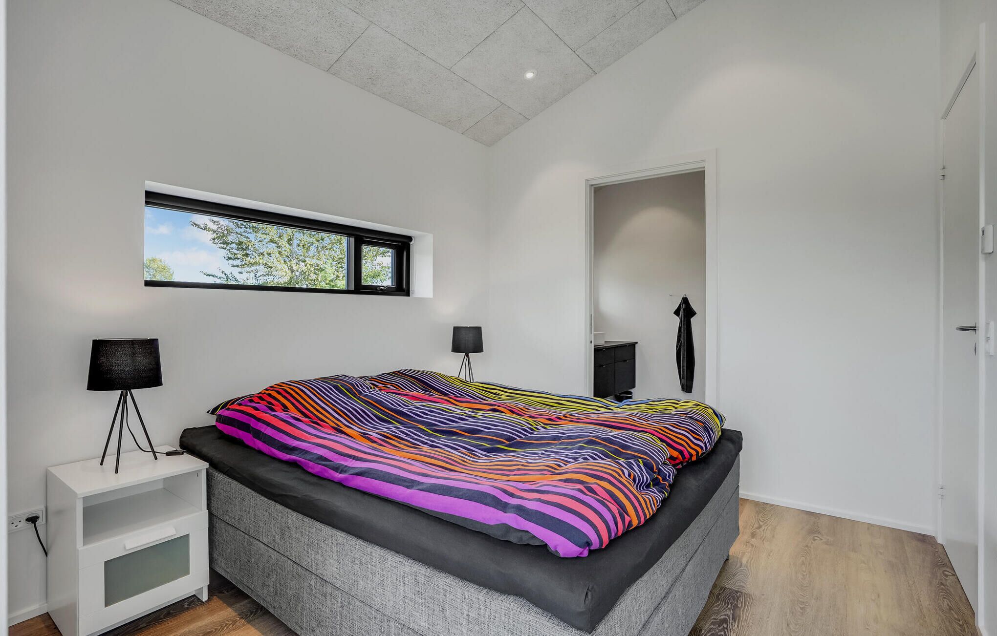 A modern bedroom with a large bed, a small bedside table, a floor lamp, and a window with a view of trees.