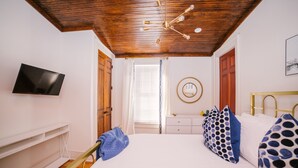 A bedroom with a wooden ceiling, a bed with white bedding, a TV on the wall, and a desk with a chair.
