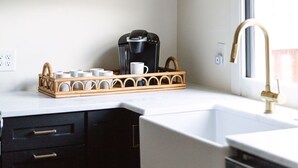 Cocina con encimeras blancas, estantes de madera, cafetera y ventana con cortina.