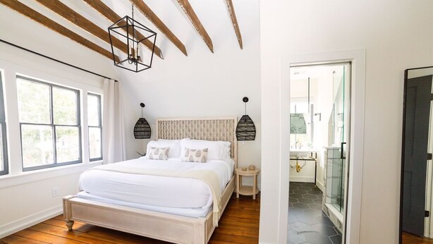 A bedroom with a wooden floor, a bed with white bedding, a headboard with a patterned design, and a hanging light fixture. - Guesthouse Charleston: EAST (46 H) (Charleston)