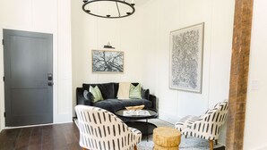 Een moderne woonkamer met een donkergrijze deur, een zwarte bank met groene en witte kussens, twee gestreepeerde fauteuils, een ronde houten salontafel en een groot botanisch kunstwerk in een lijst aan de muur.