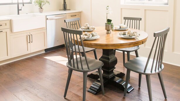 Een eetruimte met een ronde houten tafel, vier stoelen, een kroonluchter, een raam en een keuken met een spoelbak en planken.