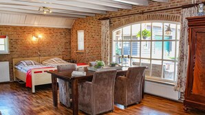 A cozy room with a wooden bed, a dining table with chairs, a large window with curtains, and a brick wall. - 1 bedroom cozy home in Udenhout (Udenhout)