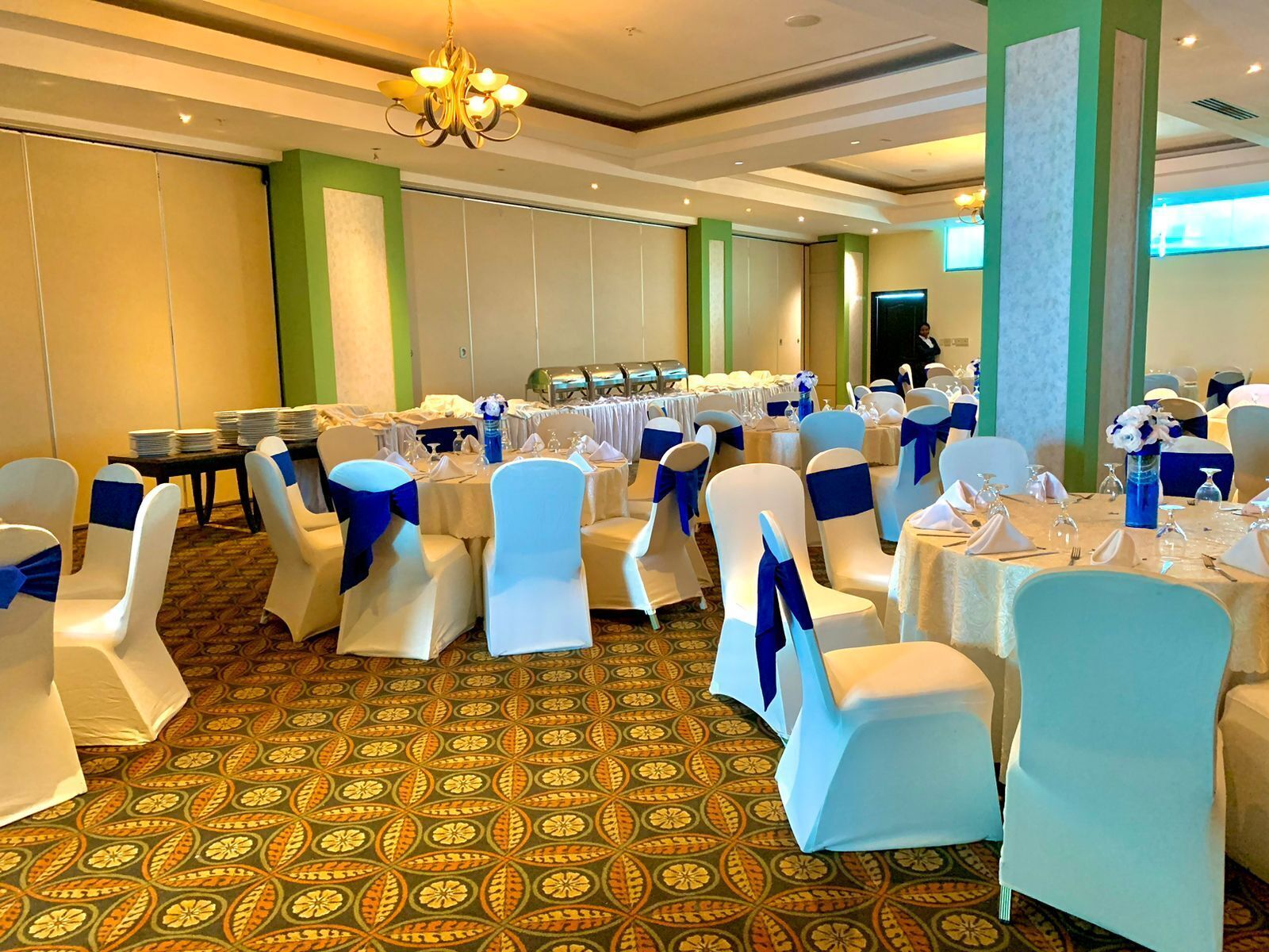 A banquet hall with round tables, white chairs with blue sashes, and a patterned carpet.