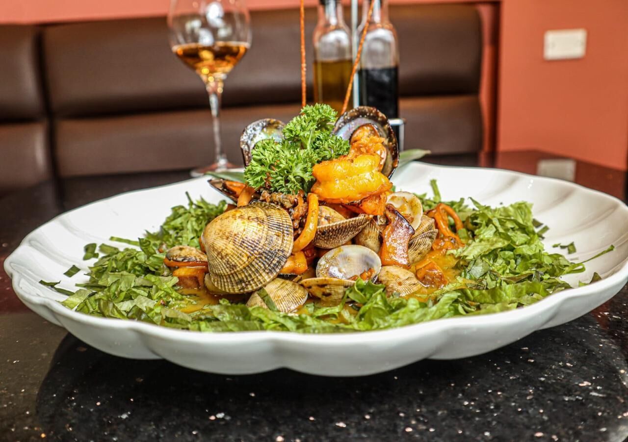 Un plato de mariscos sobre una vajilla blanca, acompañado de una copa de vino y dos botellas al fondo.