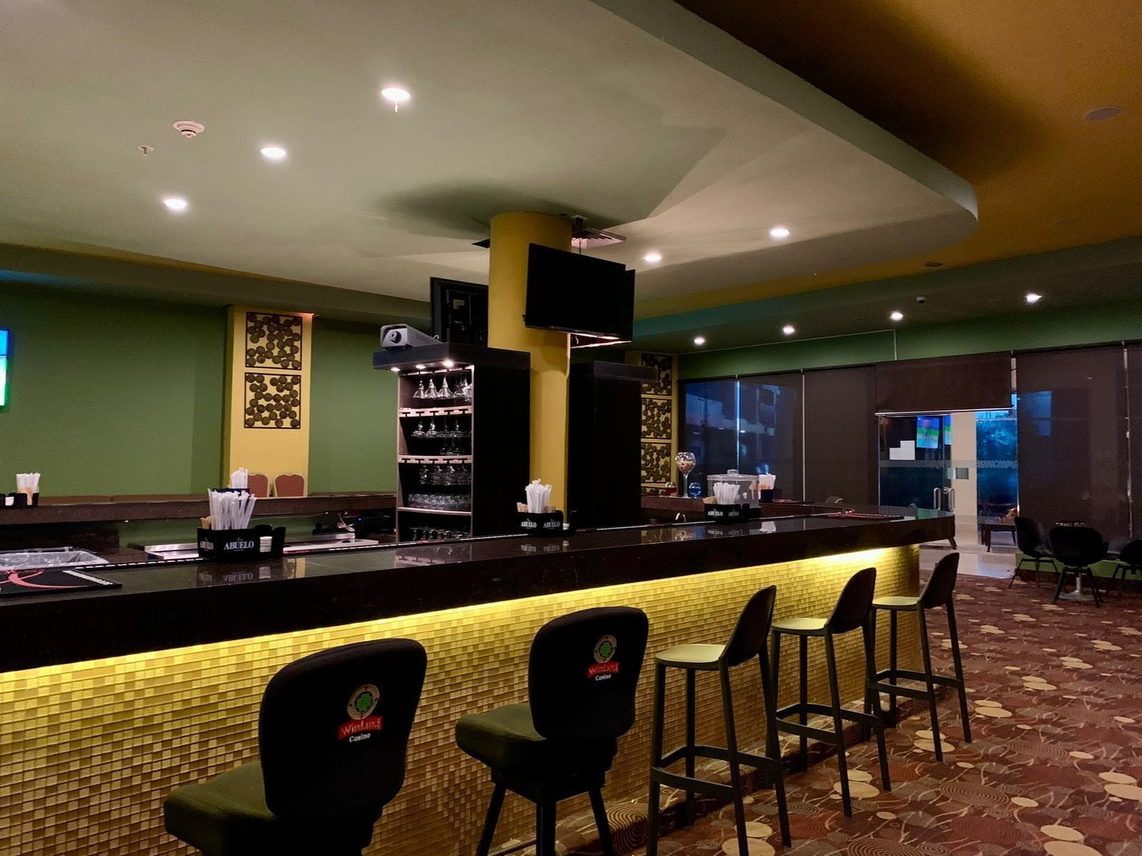 Una zona de bar con mostrador, taburetes y estanterías llenas de vasos.