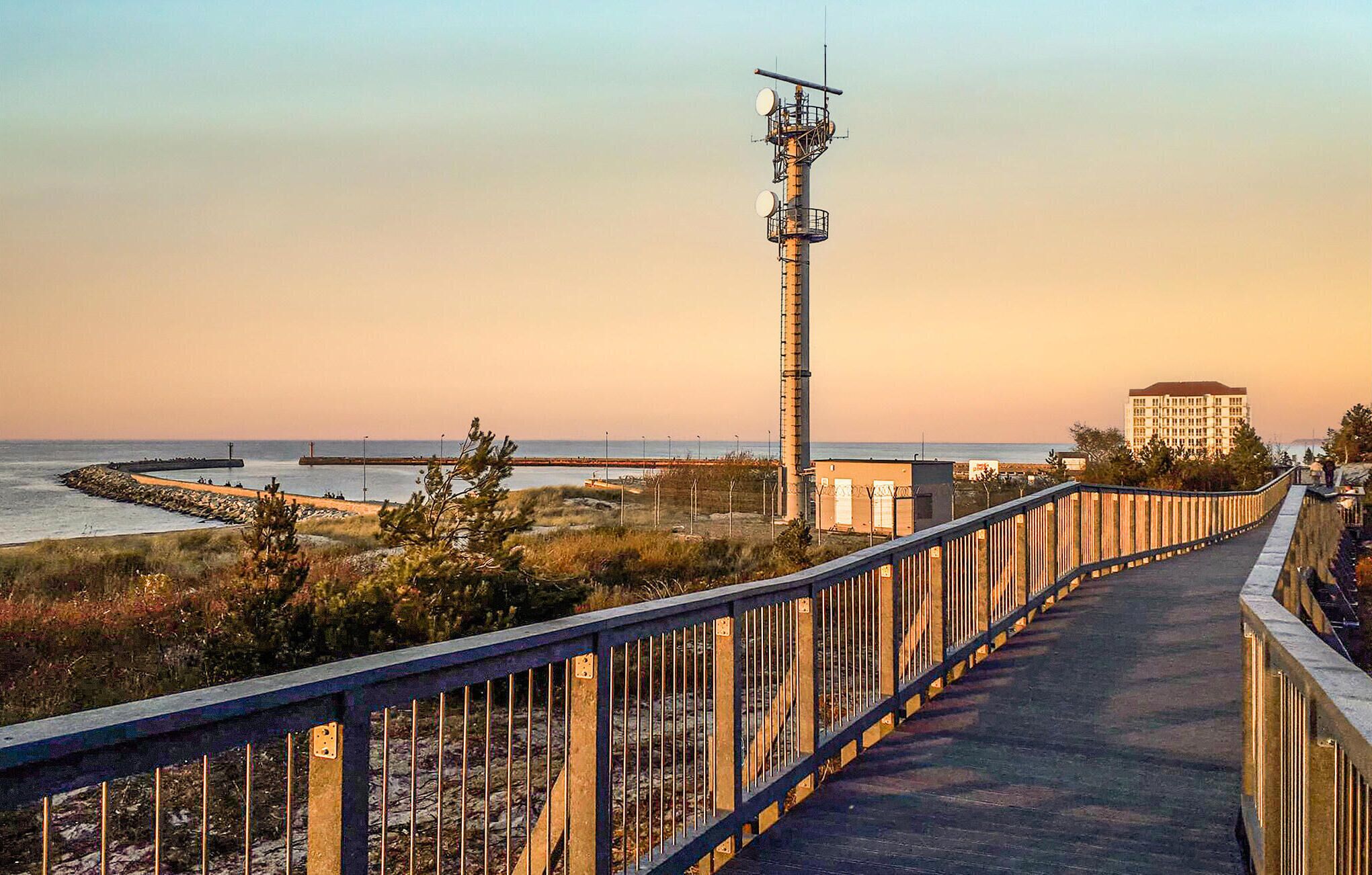 Ein Küstenweg mit Metallgeländer, einem hohen Sendeturm und einem Gebäude im Hintergrund.
