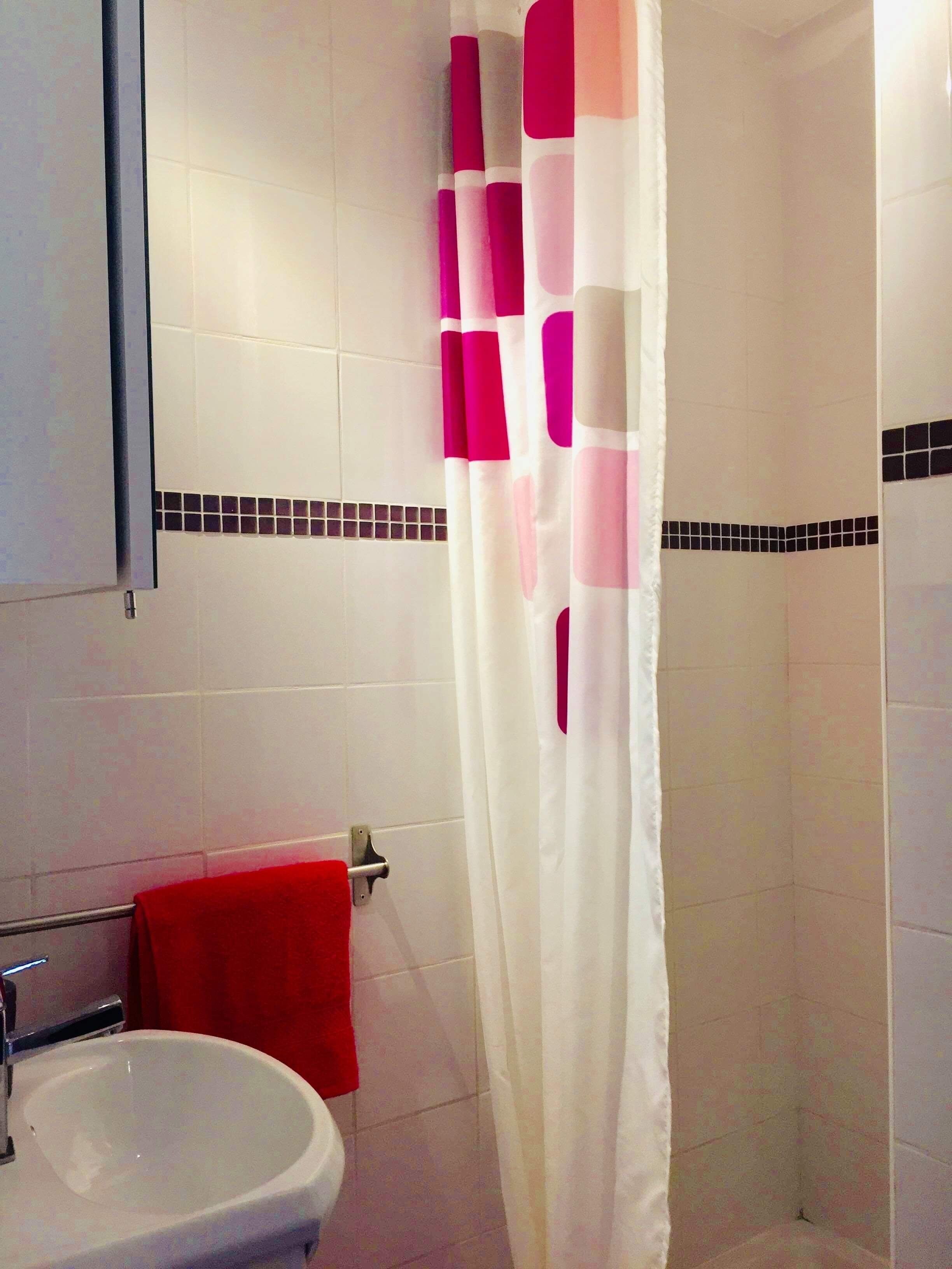 Un baño con un lavamanos blanco, una toalla roja y una cortina de ducha con motivos rosa y blanco.