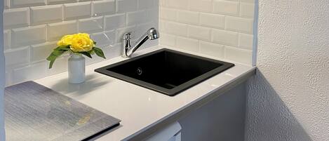 Uma cozinha com bancada branca, pia preta e um vaso branco com flores amarelas.
