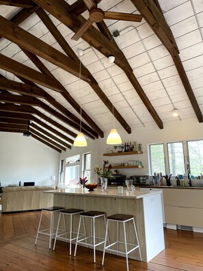 Une cuisine spacieuse dotée d’un grand îlot central, de poutres en bois, de luminaires suspendus et offrant une vue sur l’extérieur.
