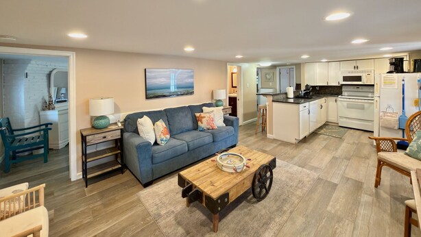 A modern living room with a blue sofa, wooden coffee table, and a kitchen area with white cabinets and appliances.