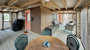 A covered outdoor area with wooden beams, a round table, wicker chairs, and a barbecue grill.