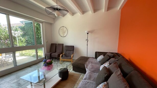 A modern living room with a glass coffee table, a dark sofa, and two armchairs. A large window provides a view of greenery outside. - Rent House Puyôo 8 people (Puyoo)