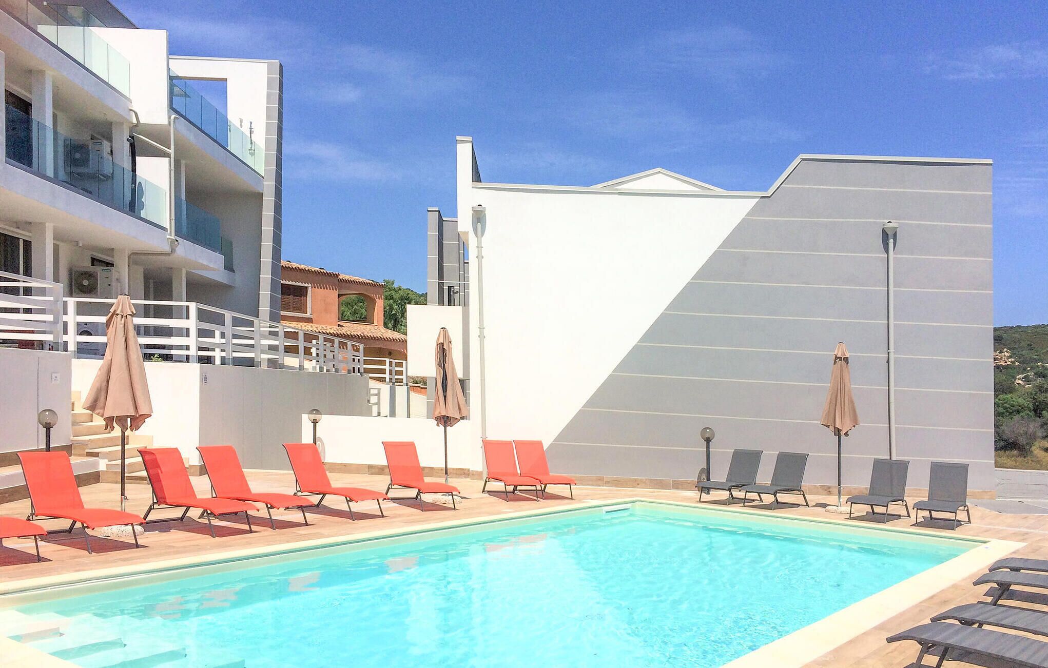 En swimmingpool med røde og sorte liggestole, en hvid bygning med en grå facade og en klar blå himmel.