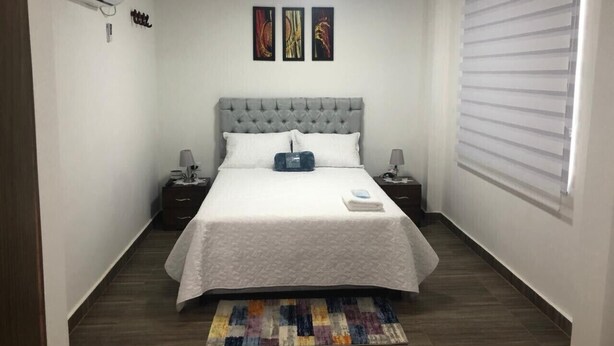 A bedroom with a bed, bedside tables, an air conditioner, artwork on the wall, and a window with blinds. - Hostal Lucy (Valledupar)