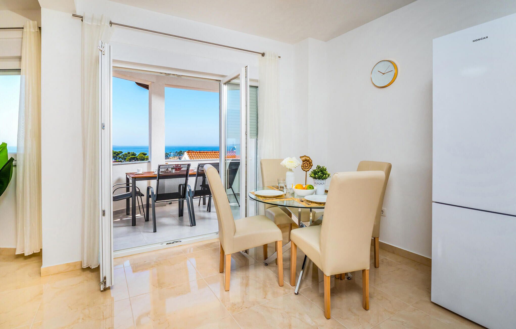 A modern dining area with a glass table, beige chairs, and a view of the sea through large windows.