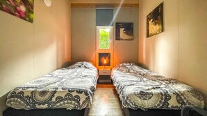 Habitación pequeña con dos camas individuales, un ventanal con vistas a zonas verdes, y una lámpara fijada a la pared.