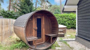 Un hidromasaje de madera con forma de barril, con puerta y escalones, rodeado por una valla de madera y follaje.