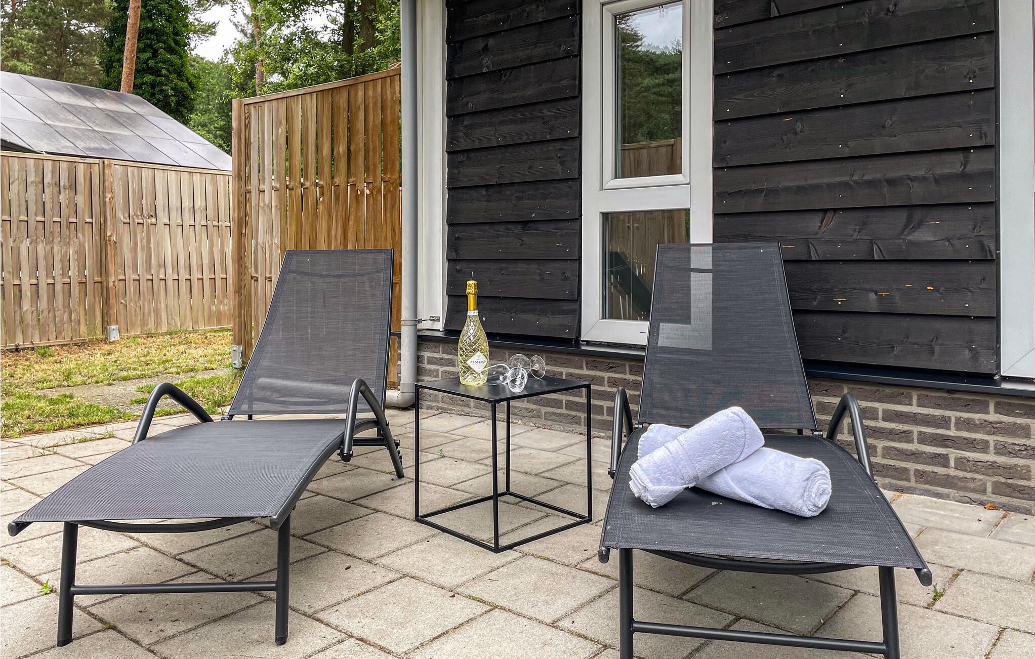 A patio with two lounge chairs and a small table, a bottle of champagne, and a rolled towel.
