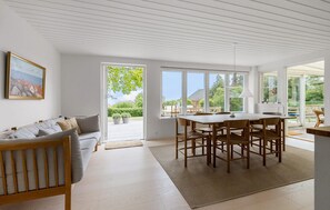 A spacious living area with a sofa, dining table, and chairs, large windows offering a view of the outdoors.