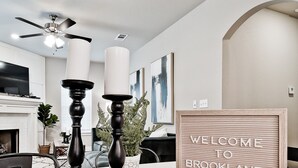 A modern living room with a round wooden table, two black candle holders, a ceiling fan, and a welcome sign.