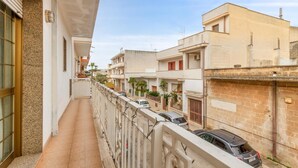 Un balcón estrecho con suelo de baldosas, barandilla metálica y vistas a edificios vecinos con coches aparcados y zonas verdes.