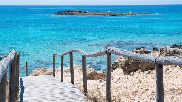 Una pasarela de madera que conduce a una costa rocosa con agua azul cristalina.