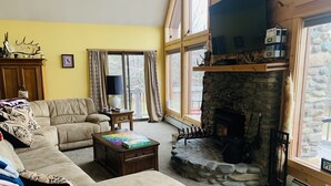 A cozy living room with a stone fireplace, a large window, and a comfortable sofa. - Spacious 3-Level Cabin Retreat-Sleeps 10, Wrap Around Deck, Peaceful and Private (Pittsburg)