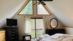 A bedroom with a sloped ceiling, a bed, a dresser, a television, a mirror, and a window with curtains. - Spacious 3-Level Cabin Retreat-Sleeps 10, Wrap Around Deck, Peaceful and Private (Pittsburg)