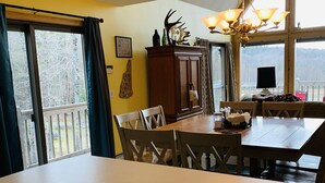 A dining area with a wooden table and chairs, a chandelier, and a view of the outdoors through large windows. - Spacious 3-Level Cabin Retreat-Sleeps 10, Wrap Around Deck, Peaceful and Private (Pittsburg)