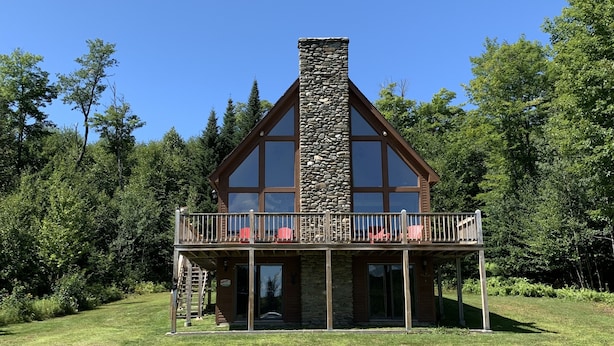 A two-story house with a stone chimney, large windows, and a balcony with red chairs. - Spacious 3-Level Cabin Retreat-Sleeps 10, Wrap Around Deck, Peaceful and Private (Pittsburg)