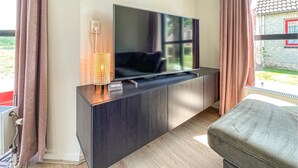Un salon moderne avec un téléviseur à écran plat posé sur un meuble en bois foncé, une petite table avec une bougie et une vue sur une maison par la fenêtre.