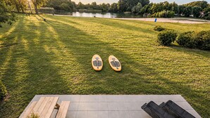 Eine grüne Außenanlage mit zwei Stand-Up-Paddles, einer Holzbank und Blick auf einen See und Bäume.