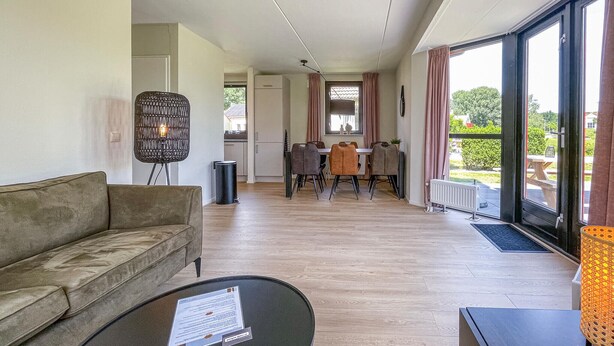 Un salon moderne comprenant un canapé, une table basse avec un avis affiché, un coin repas avec des chaises et une table, et une grande fenêtre donnant sur l’extérieur.