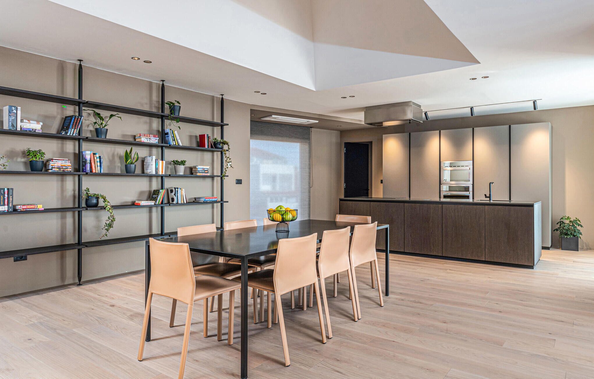 Una cocina moderna con electrodomésticos integrados, un comedor con mesa oscura y sillas claras, y una estantería con libros y plantas.