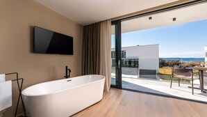 Ein modernes Badezimmer mit einer freistehenden Badewanne, einem an der Wand montierten Flachbildfernseher und einer Glastür, die auf einen Balkon mit Meerblick führt.