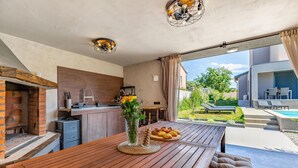 Una cucina esterna moderna con forno a mattoni, tavolo di legno e vista su piscina e giardino.