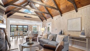 Un salon spacieux avec un plafond haut, des poutres en bois, un canapé gris, une table basse et un coin repas baigné de lumière naturelle.