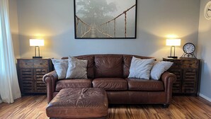 Een bruine leren bank met bijpassend voetenbankje, twee bijzettafeltjes met lampen, een lijst met een foto van een brug, en een klok aan de muur.
