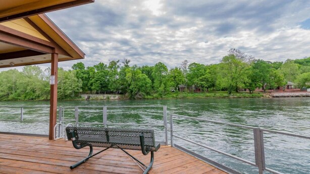 Een houten terras met een metalen bankje met uitzicht op een rivier, omringd door bomen en een bewolkte lucht.