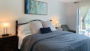 Een netjes opgemaakt bed met een grijze deken, twee bedlampen, een nachtkastje met een lamp, een klein nachtkastje met een pak tissues, en een raam met dunne gordijnen.
