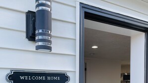 Exterior de casa com um letreiro 'Welcome Home', uma câmera de segurança e uma porta que dá para uma sala de jantar com mesa e cadeiras.