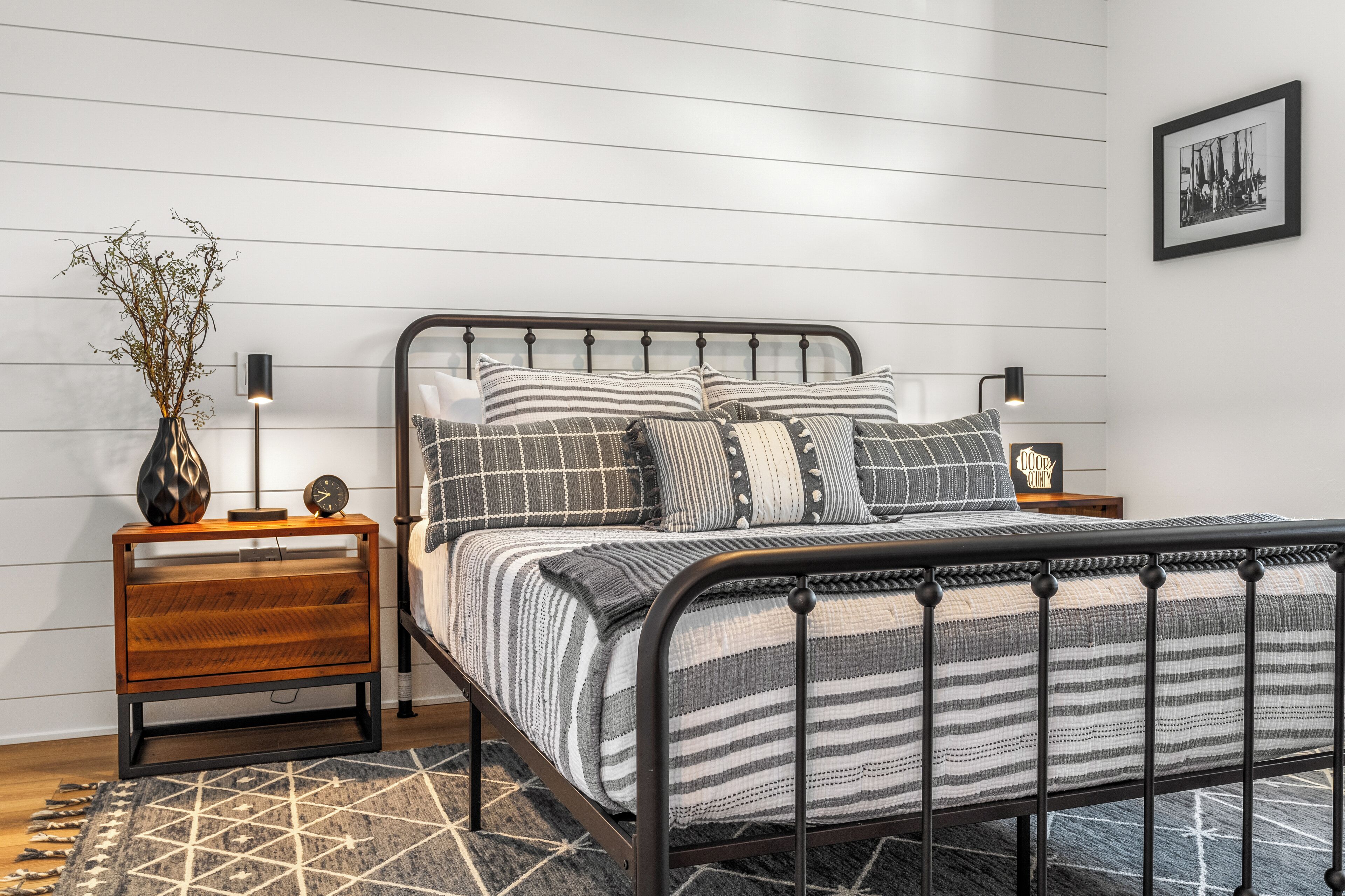 A modern bedroom with a metal bed frame, a wooden nightstand with drawers, a bedside table, a wall-mounted lamp, and a framed picture on the wall.