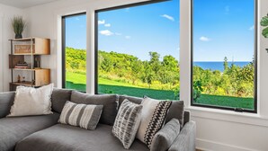 Una sala moderna con un sofá grisáceo, cojines estampados y una estantería. Amplias ventanas ofrecen vistas a un paisaje verde exuberante y al océano.