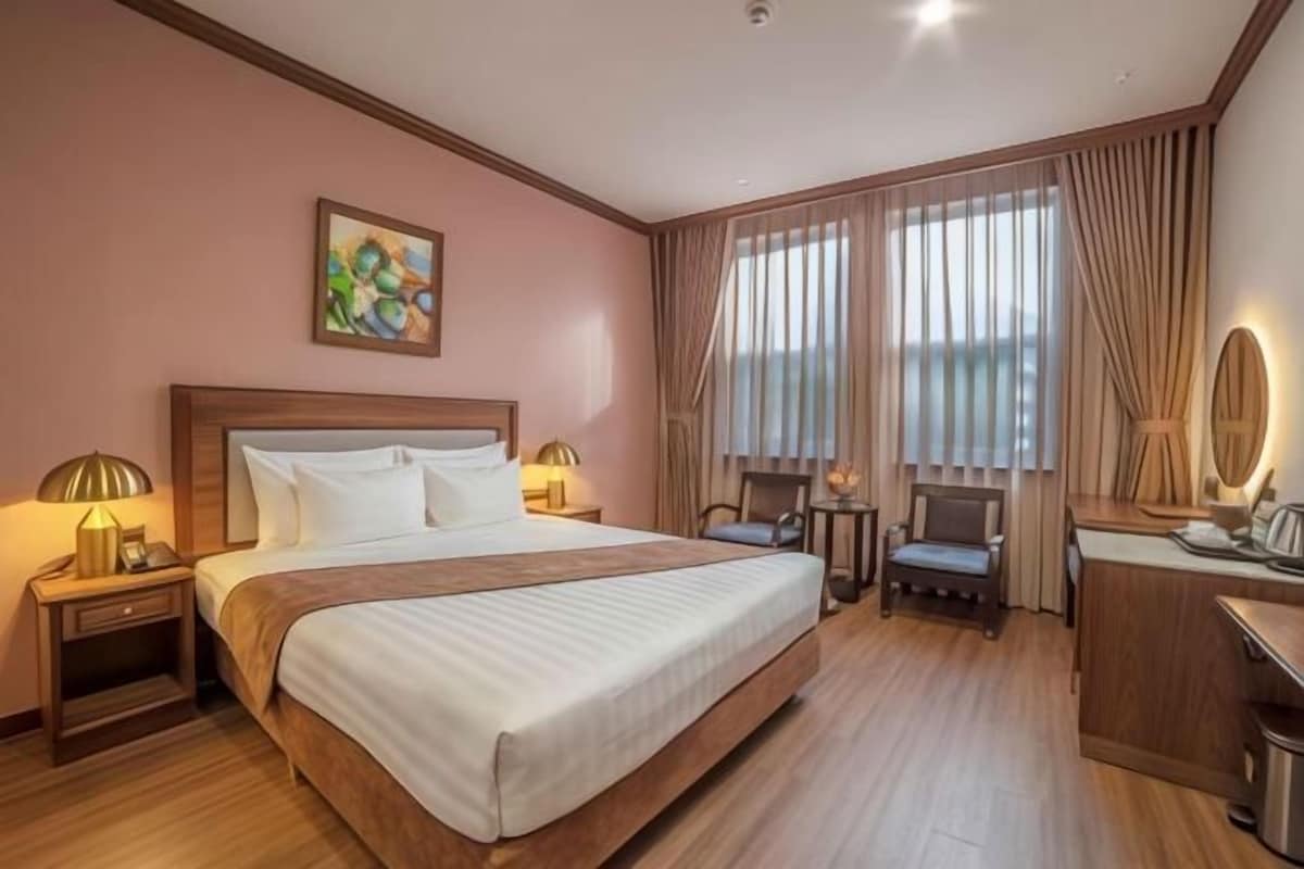 Habitación de hotel con una cama grande, dos mesitas de noche con lámparas, un escritorio con una silla, una pequeña mesa con una lámpara y un amplio ventanal con cortinas.