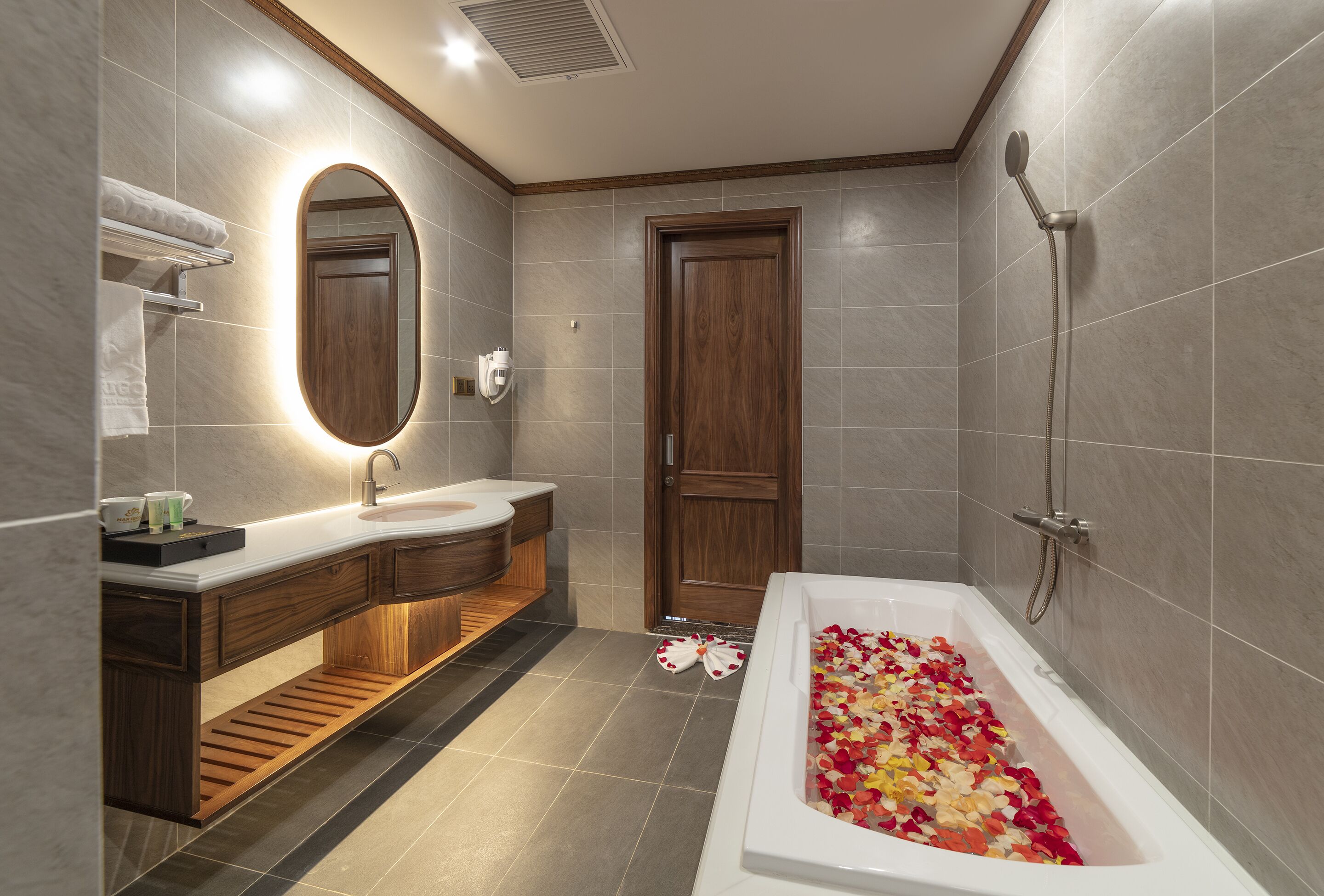 A modern bathroom with a wooden vanity, a large mirror, a bathtub filled with rose petals, and a shower area.